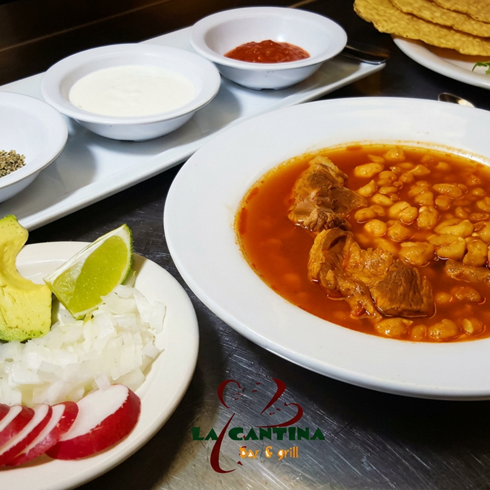 Mexican pozole with pork and hominy served alongside avocado, lime, onion, radishes, spices, sauces, and tostadas.