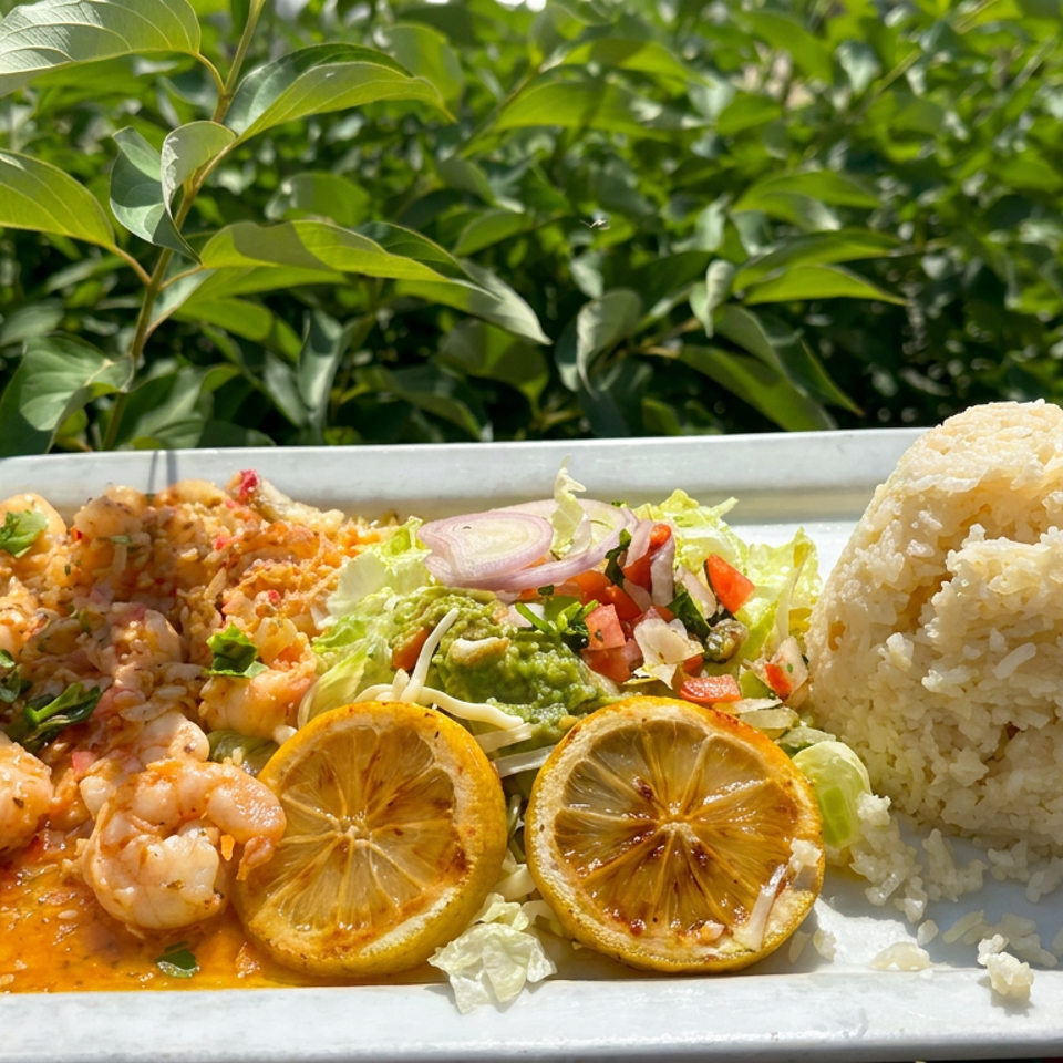 Plate of saucy shrimp, molded white rice, salad, guacamole, and grilled lemon slices served outdoors amid lush greenery.