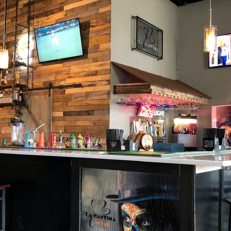 Industrial-style La Cantina restaurant bar with pendant lights, TVs, colorful artwork, beverage taps, and high-top seating.