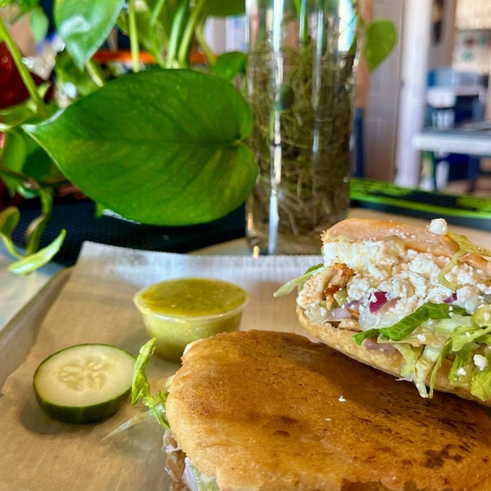 Loaded sandwich with crispy filling, lettuce, and creamy sauce on a wooden board, accompanied by cucumber slices and green dipping sauce.
